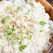 White coconut rice in a wooden bowl. The rice is topped with coconut flakes and chopped green herbs with a lime wedge on the side.
