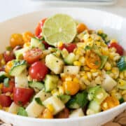 A corn salad with zucchini, cherry tomatoes, and fresh herbs in a white bowl.