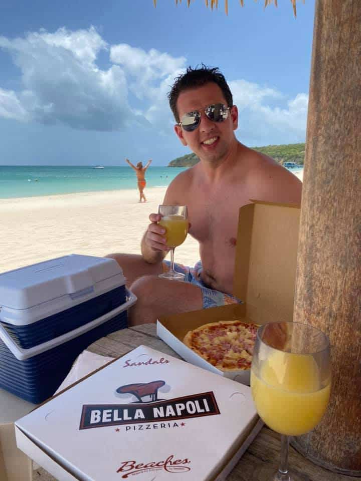 jj sitting on the beach at Sandals Grande Antigua holding a mimosa with pizza. Travel Guide.