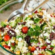 A fresh couscous salad with vegetables and olives in a clear bowl with two spoons digging in.