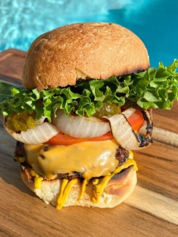 Grilled cheeseburger with all the toppings on a cutting board in front of a pool. Beef burger with onions, tomatoes, pickles, lettuce, and mustard.