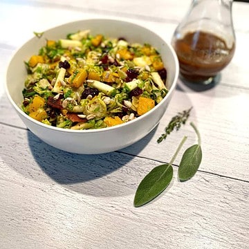 bowl of Butternut Squash and Brussels Sprouts Salad with sage and thyme leaves on the table. Maple balsamic salad dressing is in a jar to the side of the salad bowl.
