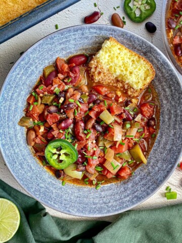 Three bean chili in a grey bowl. A pan of cornbread is next to the bowl along with beer caps, sliced jalapenos, and beans.