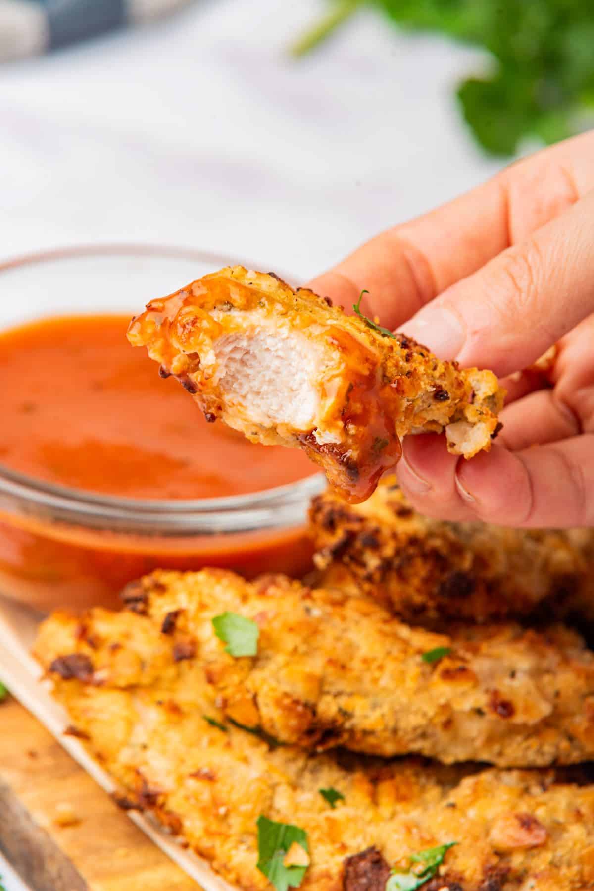 A hand holding crispy breaded cheesy chicken with a bite taken out of it to show the juicy white center.