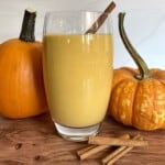 Orange colored pumpkin pie smoothie in a glass with real pumpkins on either side of the glass. Cinnamon sticks are next to the glass.