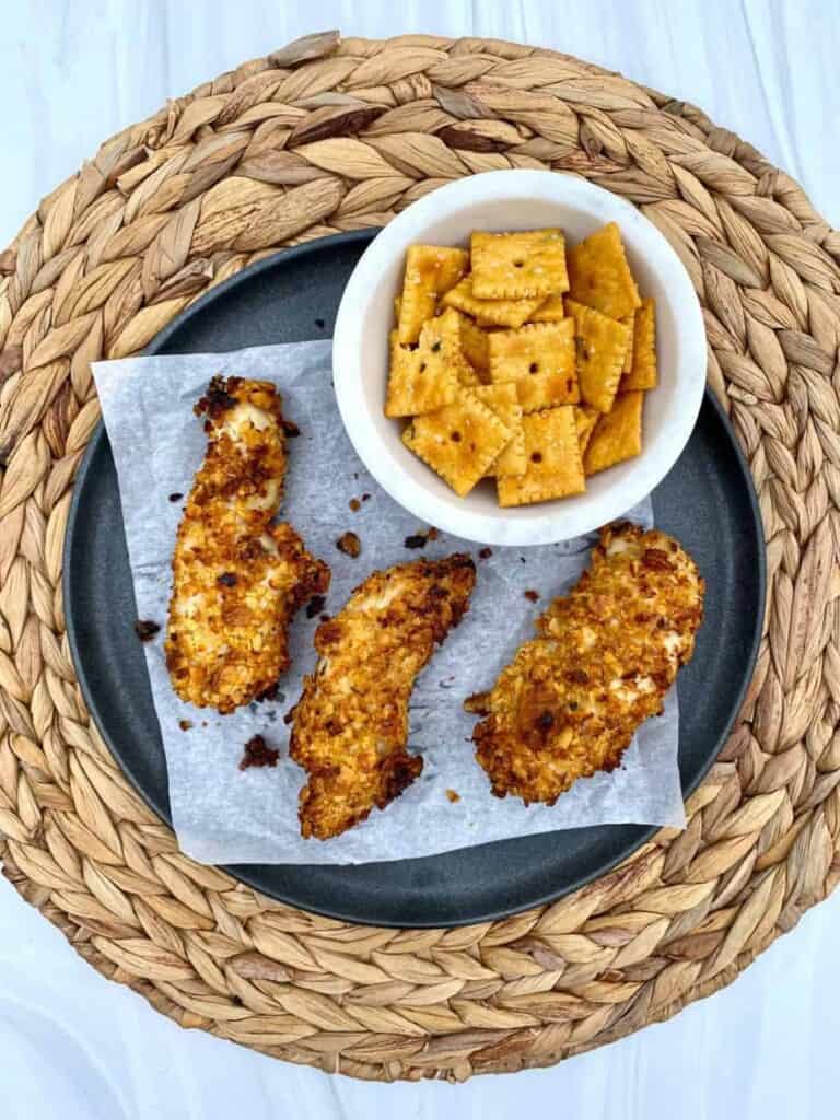Three Air Fryer Cheez It chicken tenders on a plate with a bowl of cheez it crackers on the side.