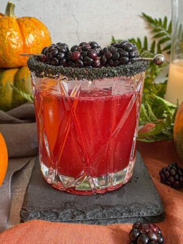 Dark and Stormy Halloween cocktail with whiskey. The glass is garnished with a black sugar rim and three blackberries on a silver skewer. Fresh pumpkins and a candle is lit in the background.