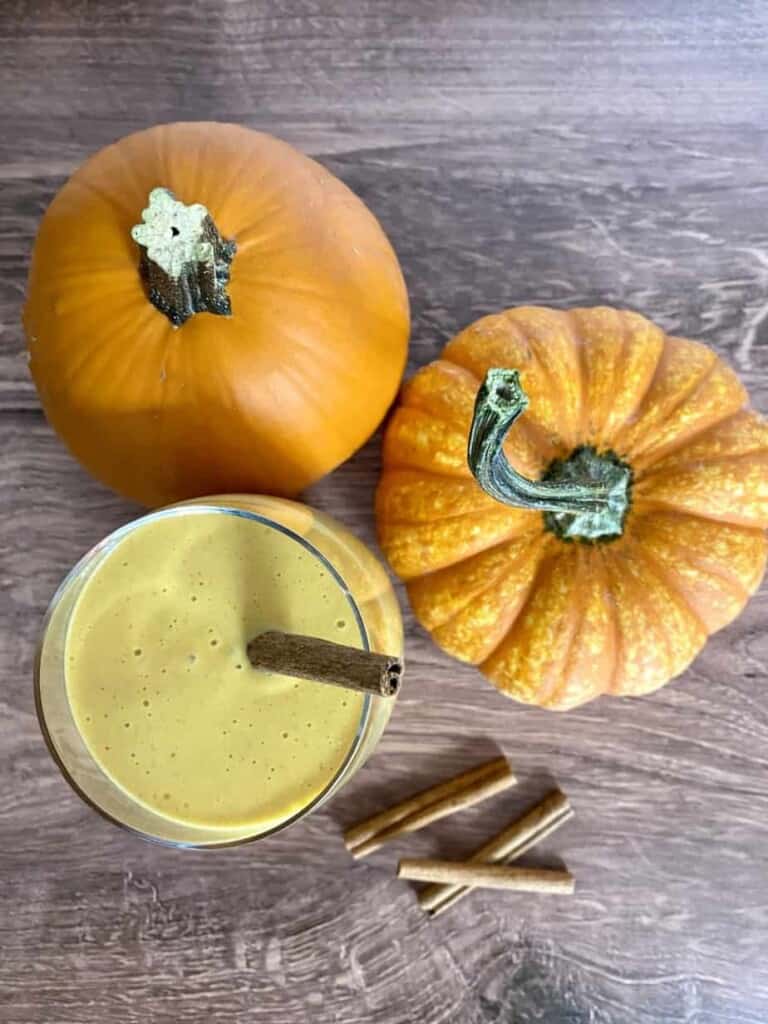 Looking down on a glass filled with pumpkin spice smoothie. Two orange pumpkins and cinnamon sticks are on a wooden table with the glass.