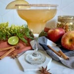 Thanksgiving margarita in a a margarita glass and an apple slice as a garnish. Apple, a sliced lime, and greenery surround the cocktail.