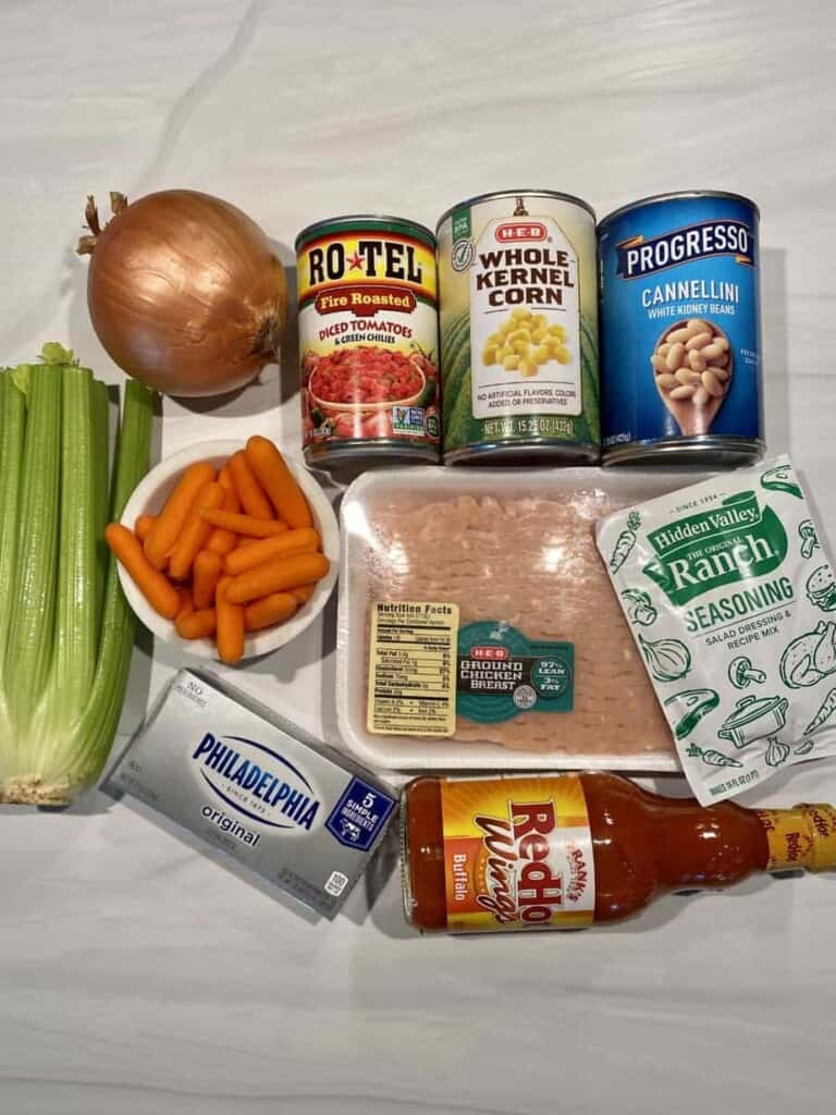 Fresh and canned ingredients needed to make this Creamy Buffalo Chicken soup recipe are on a table: ground chicken, onion, celery, carrots, cream cheese, beans, and buffalo sauce.