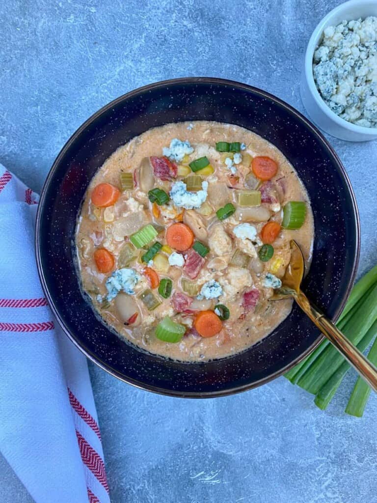 A bowl of buffalo chicken soup with scallions and blue cheese on the side. The soup is full of vegetables, such as carrots, celery, onions, and tomatoes.