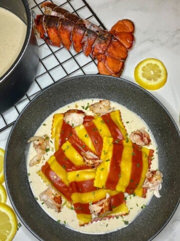 Colorful lobster ravioli sits in a bowl with sherry cream sauce. A lobster tail, lemon slices, and gold fork are next to the bowl of food.