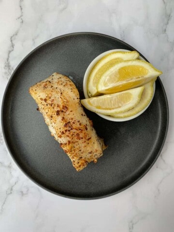 Air fried Chilean sea bass fillet on a plate with lemon wedges.