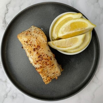 Air fried Chilean sea bass fillet on a plate with lemon wedges.