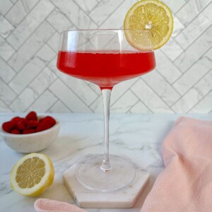 A red raspberry elderflower gin cocktail in a coupe glass with a lemon slice as a garnish.