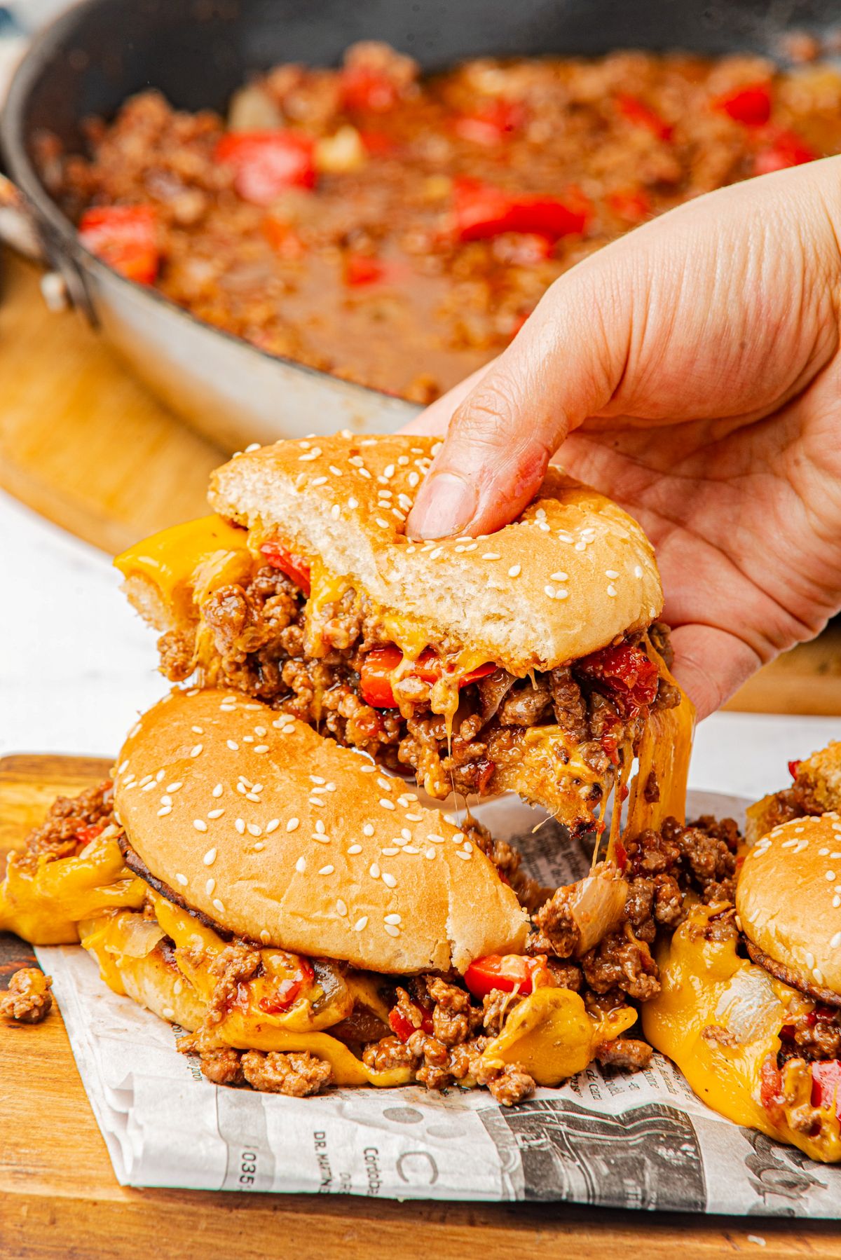 A hand pulling apart a cheesy sloppy joes sandwich next to the homemade sauce in a skillet.