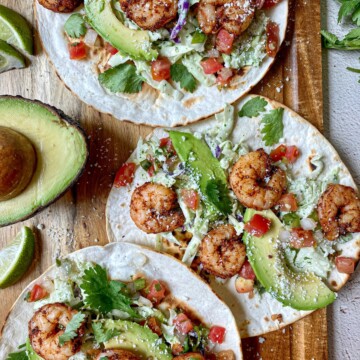 Three air fried shrimp tacos with slaw, avocado, cilantro, and creamy jalapeno salsa are open face on a cutting board. Lime wedges and an avocado are on the side.