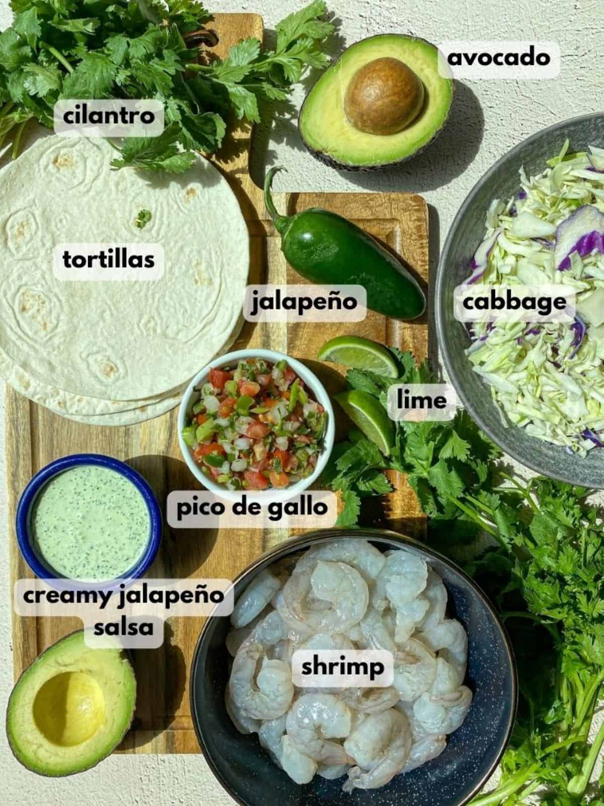 Shrimp and cabbage in a bowl, a jalapeno, tortillas, halved avocado, and cilantro are on a wooden cutting board.