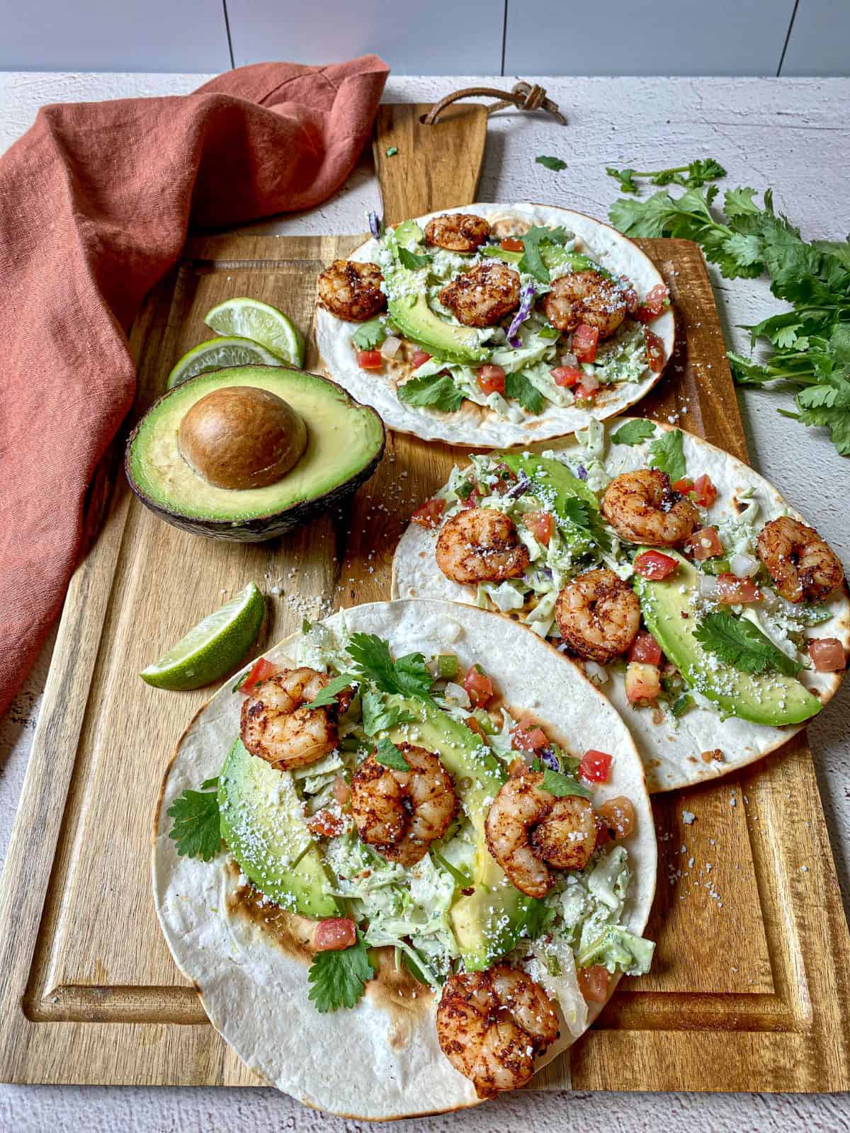 Three air fryer shrimp tacos with slaw on a cutting board with an avocado, cilantro, and lime wedges.