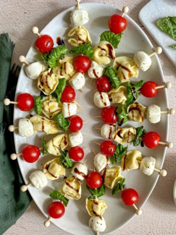 Caprese tortellini skewers with balsamic glaze on a serving platter. Cherry tomatoes, fresh basil, and a green kitchen towel surround the plate.