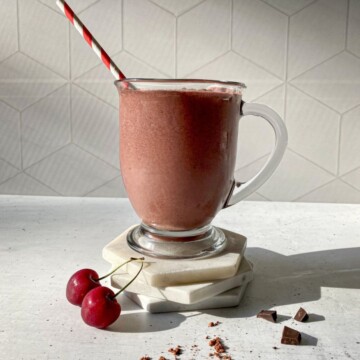 Chocolate Cherry Smoothie in a glass with a straw. Cherries, a dusting of cacao powder, and chocolate chucks are around the glass.