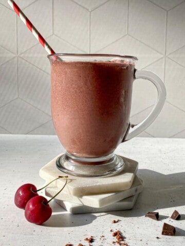 Chocolate Cherry Smoothie in a glass with a straw. Cherries, a dusting of cacao powder, and chocolate chucks are around the glass.
