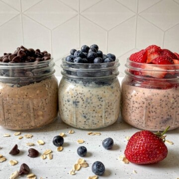 Three mason jars filled with protein overnight oats. The three flavors include: Chocolate chip overnight oats have chocolate chips on top, blueberry cheesecake overnight oats has blueberries on tope, and strawberry shortcake overnight oats has chopped strawberries on top.