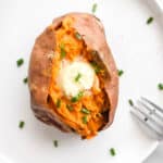 A whole air fryer sweet potato with butter and chives on the top. A fork is on the side of the plate.