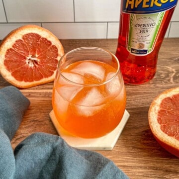 A glass of an aperol gin cocktail. It's an orange colored cocktail with gin and aperol. A bottle of aperol and a sliced grapefruit are on the side of the cocktail with a blue cloth.