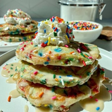 Two stacks of homemade funfetti pancakes with sprinkles on white plates with a bowl of rainbow sprinkles on the side.