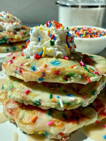 Two stacks of homemade funfetti pancakes with sprinkles on white plates with a bowl of rainbow sprinkles on the side.