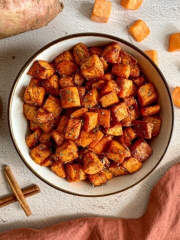 A bowl of air fried sweet potato cubes. Whole and cubed sweet potatoes, cinnamon sticks, and measuring spoons with nutmeg and ginger surround the bowl.