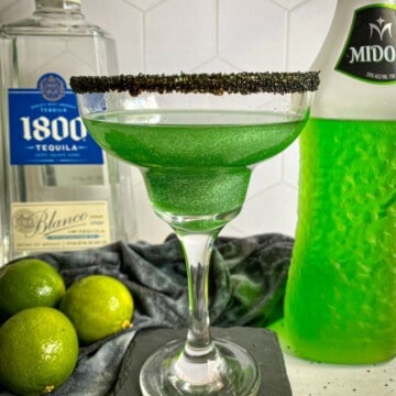 Green Halloween-themed margarita made with midori with a black sugar rim. A bottle of midori melon liqueur and tequila are in the background with three limes.