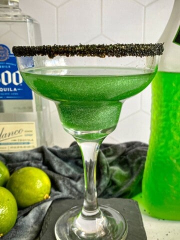 Green Halloween-themed margarita made with midori with a black sugar rim. A bottle of midori melon liqueur and tequila are in the background with three limes.