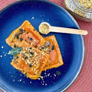 Pumpkin ravioli on a blue plate. Walnut parmesan and fried sage leaves are on top of the pasta. A jar of walnut parmesan and a measuring spoon are on the side.
