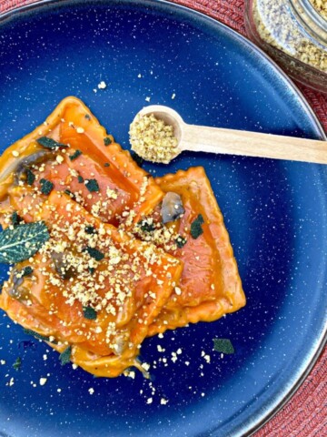 Pumpkin ravioli on a blue plate. Walnut parmesan and fried sage leaves are on top of the pasta. A jar of walnut parmesan and a measuring spoon are on the side.