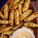 Crispy potato wedges are on a wooden plate with a small bowl of dipping sauce to the side.