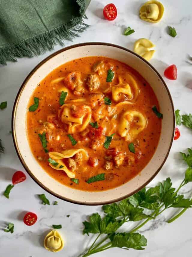 A bowl of Tomato Tortellini Soup with Italian Sausage. Tortellini and tomatoes surround the beige bowl of soup.