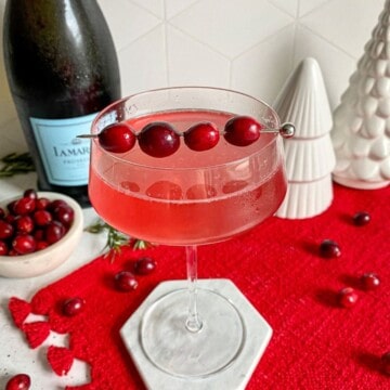 Cranberry gin cocktail in a coupe glass with a garnish of fresh cranberries on top of a red placemat. A bottle of prosecco and fake white Christmas tree decorations are in the background.