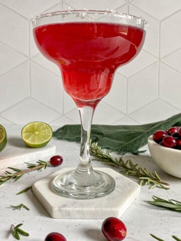 A cranberry margarita in a glass with a salt rim surrounded by fresh cranberries and limes.