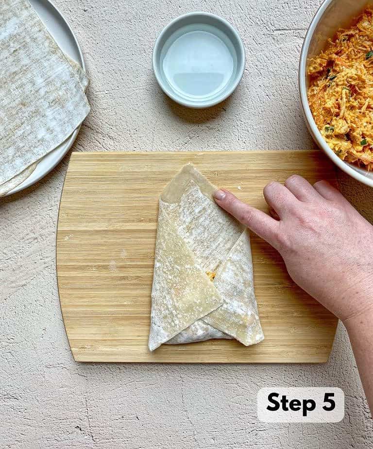 A finger shown rubbing water on the top corner of the egg roll wrapper to help seal tightly.