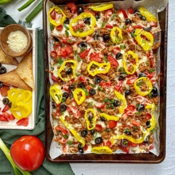 Italian Nachos on a sheet pan topped with sausage, alfredo sauce, cheese, tomatoes, olives, and peppers. A tray with extra toppings and grated cheese is next to the pan.