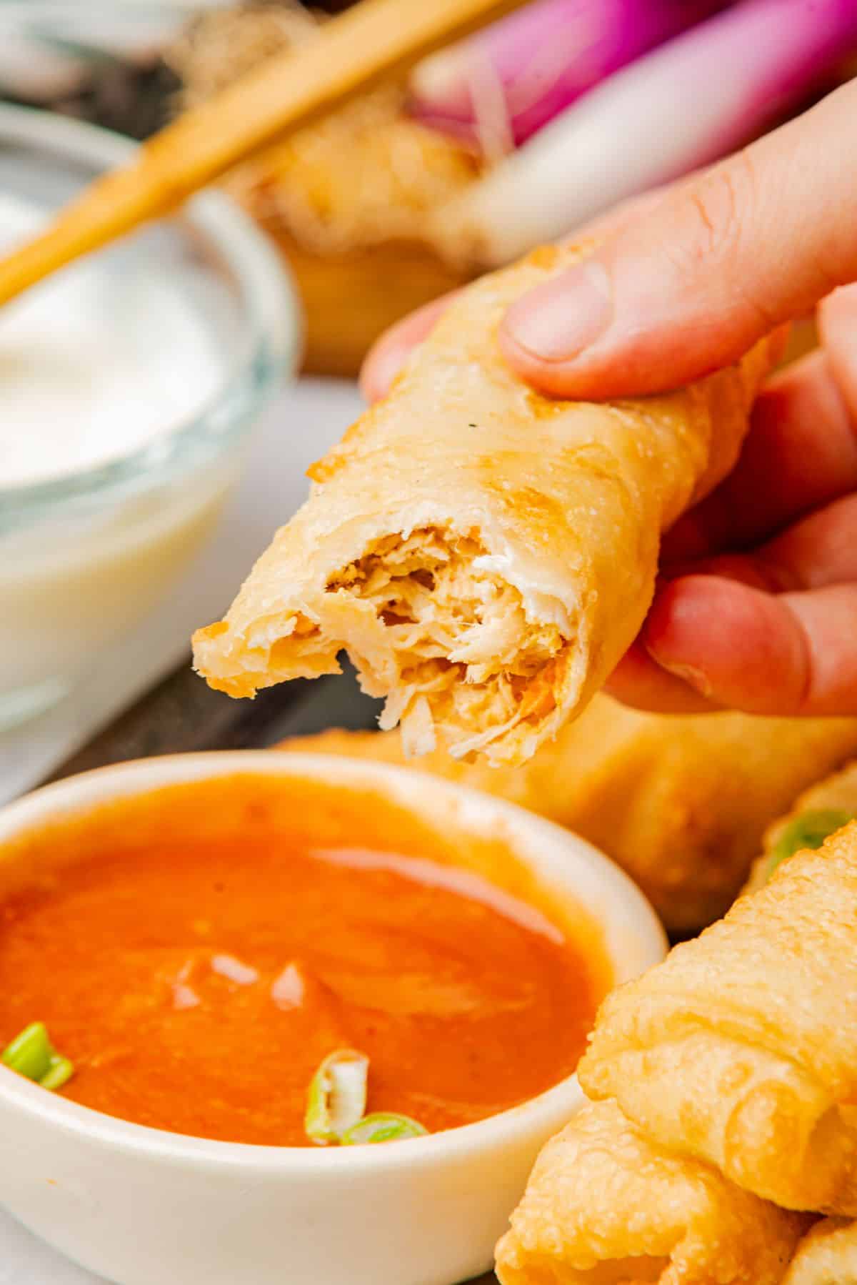 A hand holding a buffalo chicken egg roll with a bite taken out to show the mixture on the interior.