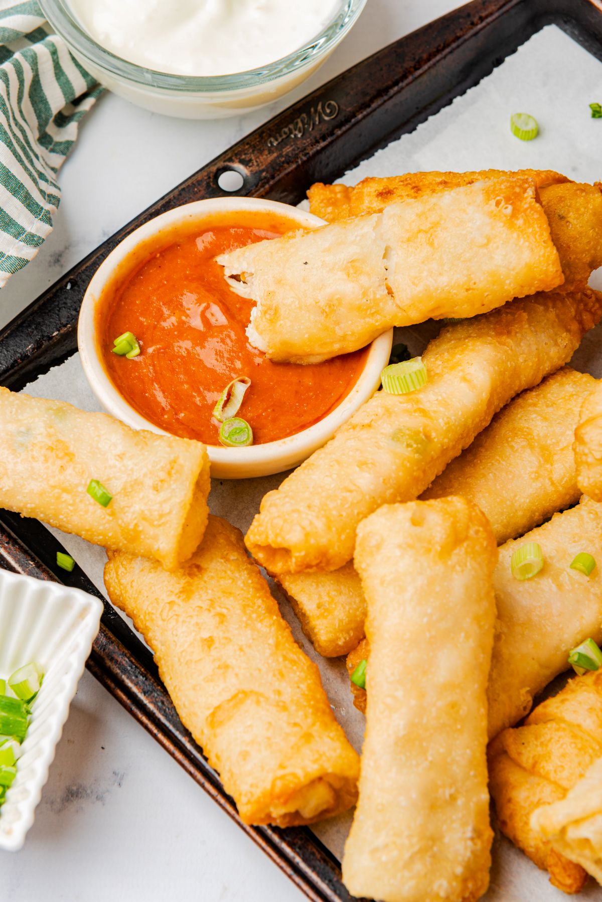 Buffalo chicken egg rolls on a serving platter with one dripping into a bowl of buffalo sauce.