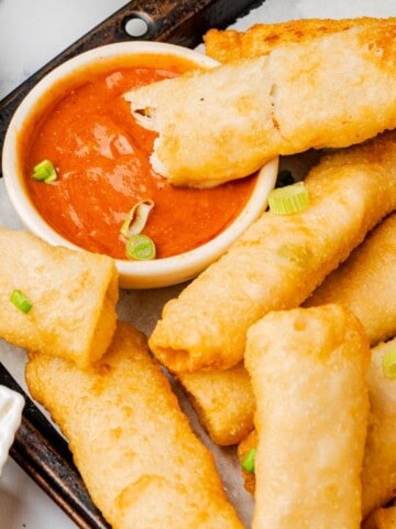 Buffalo chicken egg rolls on a serving platter with one dripping into a bowl of buffalo sauce.