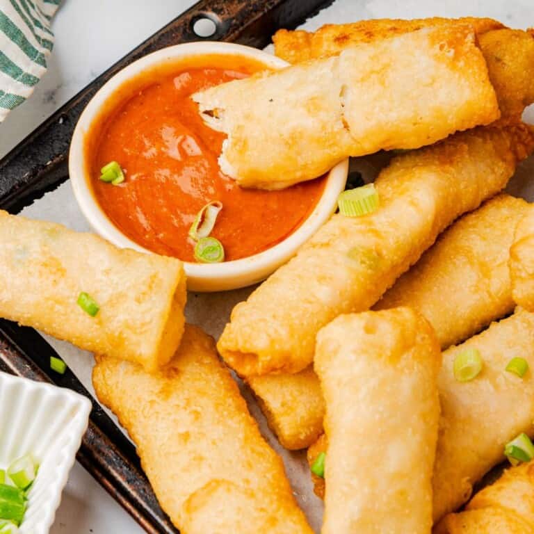 Buffalo chicken egg rolls on a serving platter with one dripping into a bowl of buffalo sauce.