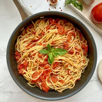 Spaghetti Arrabiata, in a skillet with red pepper flakes, garlic cloves, basil, and a roma tomato on the side.