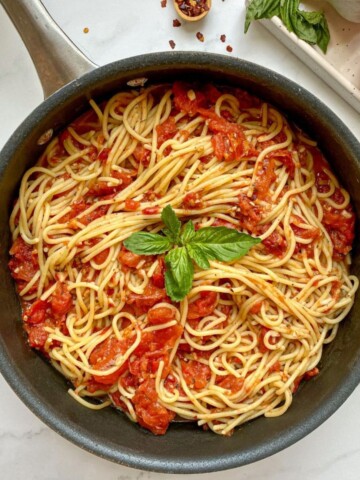 Spaghetti Arrabiata, in a skillet with red pepper flakes, garlic cloves, basil, and a roma tomato on the side.