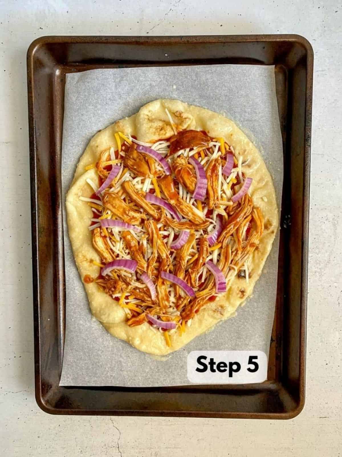 Step five. BBQ sauce, cheese, shredded chicken, and red onions on naan pizza crust that is on a baking sheet with parchment paper.