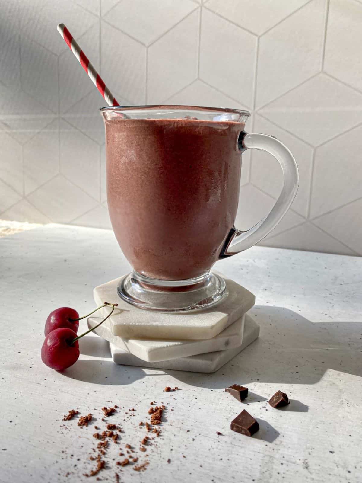 Chocolate Cherry Smoothie with Cacao in a glass with a straw. Cherries, a dusting of cacao powder, and chocolate chucks are around the glass.